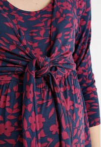 Laivastonsininen mekko, jossa on tummanpunainen kukkakuosi; edessä solmittava solmu ja kolmannesosa hihasta; valmistettu pehmeästä, joustavasta kankaasta.