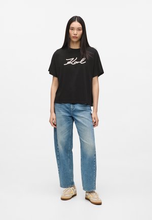 Ung person med langt mørkt hår iført sort "Karl" T-shirt, lyseblå jeans og beige sneakers, stående foran en ensfarvet hvid baggrund.