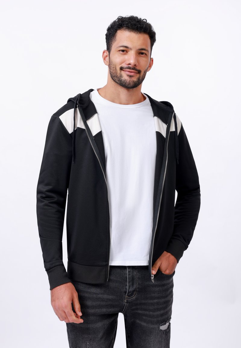 Sweat à capuche noir avec des accents blancs sur les épaules, en tissu doux. Comprend une capuche ajustable et des poignets côtelés. Portez-le par-dessus une chemise blanche.