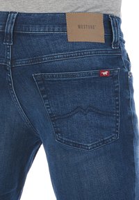 Jeans de denim azul oscuro con una etiqueta de cuero marrón que dice "MUSTANG" en la cintura y un pequeño logo rojo en el bolsillo trasero.