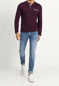 Pitkähihainen, tummanvioletti henley-paita, jossa napillinen kaulus, yhdistetty vaaleansinisiin farkkuihin ja valkoisiin lenkkareihin, kävelemässä neutraalilla taustalla.