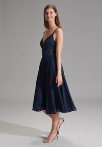 Robe bleu marine avec un corsage orné de perles, décolleté en V et jupe fluide arrivant au genou. Associée à des sandales argentées à lanières.