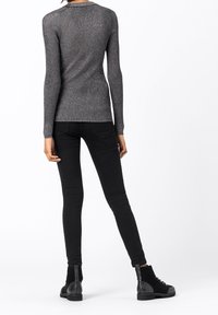 Kobieta w dopasowanej, szarej, metalicznej bluzce z długim rękawem, czarnych rurkach i czarnych, sznurowanych botkach za kostkę, stoi tyłem.