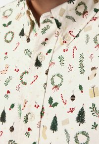 Camicia bianca con bottoni decorata con motivi natalizi tra cui corone, alberi, bastoncini di zucchero, calze, agrifoglio, bacche e scatole regalo.