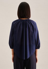 Person mit kurzem glattem Haar, die eine lockere marineblaue Bluse mit gerafftem Ausschnitt und Dreiviertelärmeln trägt, von hinten gezeigt.