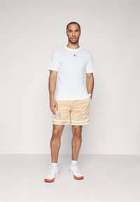 Weißes Kurzarm-T-Shirt mit schwarzem Logo, kombiniert mit beigen Mesh-Shorts mit weißen Akzenten und gestickten Aufnähern. Sportschuhe mit roten Details.