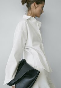 Femme vêtue d'une chemise blanche surdimensionnée et d'un pantalon, tenant un grand sac à main en cuir noir sur un fond gris clair uni.