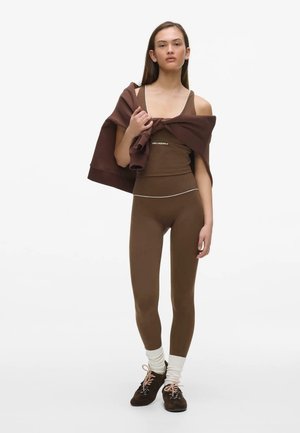 Donna con canotta marrone e leggings, calzini bianchi, scarpe da ginnastica scure, che tiene una felpa marrone appoggiata sulle spalle su sfondo bianco.