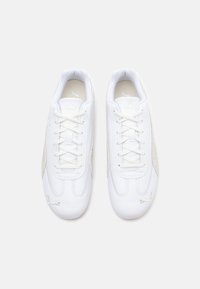 Baskets blanches en cuir lisse avec des accents en suède, présentant un design à lacets et des logos bien visibles sur le côté.