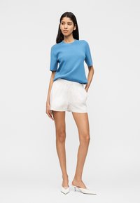 Jeune femme portant un haut bleu à manches courtes, un short blanc avec cordon de serrage et des mules blanches à talons, debout avec une main dans la poche.