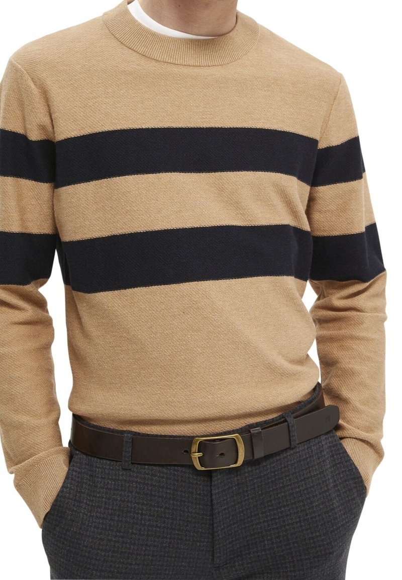 Homme portant un pull beige à rayures horizontales noires, associé à un pantalon à carreaux foncé et une ceinture en cuir marron avec une boucle en laiton.