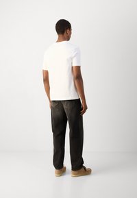 Witte korte mouwen t-shirt gecombineerd met zwarte loszittende jeans en lichtbruine schoenen, gezien vanuit de achterkant tegen een neutrale achtergrond.