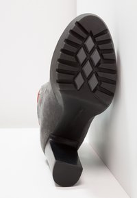 Bottine en suède gris avec un talon rectangulaire, semelle noire texturée présentant un motif en diamant, et détail en rouge sur le côté.