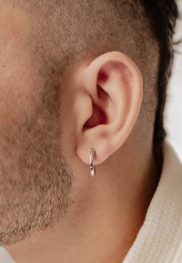 Orecchino a cerchio argentato mostrato all'orecchio di una persona. L'orecchino è liscio, circolare e riflette la luce, in contrasto con i capelli tagliati corti.