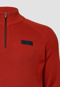 Czerwony sweter z prążkowanej dzianiny z wysokim kołnierzem, posiadający pół zamek oraz czarną łatę z logo na lewym ramieniu. Teksturowana dzianina.
