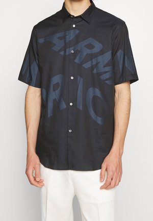 Czarna koszula na krótki rękaw zapinana na guziki z dużymi ciemnoniebieskimi abstrakcyjnymi literami, noszona przez osobę w białych spodniach na jednolitym tle.