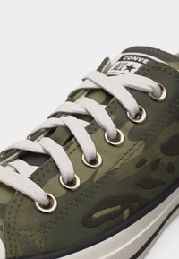 Κοντινό πλάνο σε καμβά αθλητικού παπουτσιού με χακί παραλλαγή, λευκά κορδόνια και μεταλλικά ρουξούνια, εμφανίζοντας μέρος της ετικέτας "Converse All Star".