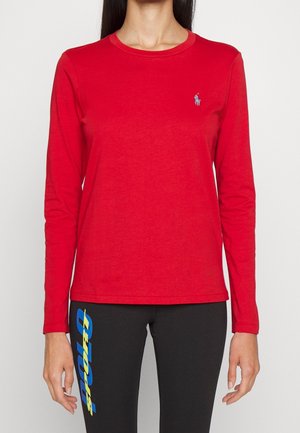 Kobieta ubrana w czerwoną bluzkę z długim rękawem z małym logo na piersi oraz czarne legginsy z pionowym niebiesko-żółtym napisem.