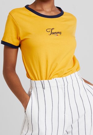 Osoba nosząca żółtą koszulkę z granatowymi obszyciami i logo "Tommy", zestawioną z białymi spodniami z wysokim stanem i pionowymi czarnymi paskami.