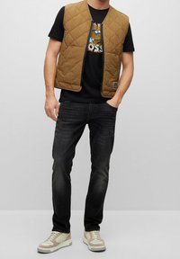 Gilet marrone trapuntato sopra una t-shirt nera con grafica, abbinato a jeans scuri e sneakers beige. Chiusura con cerniera, vestibilità casual, senza loghi visibili.