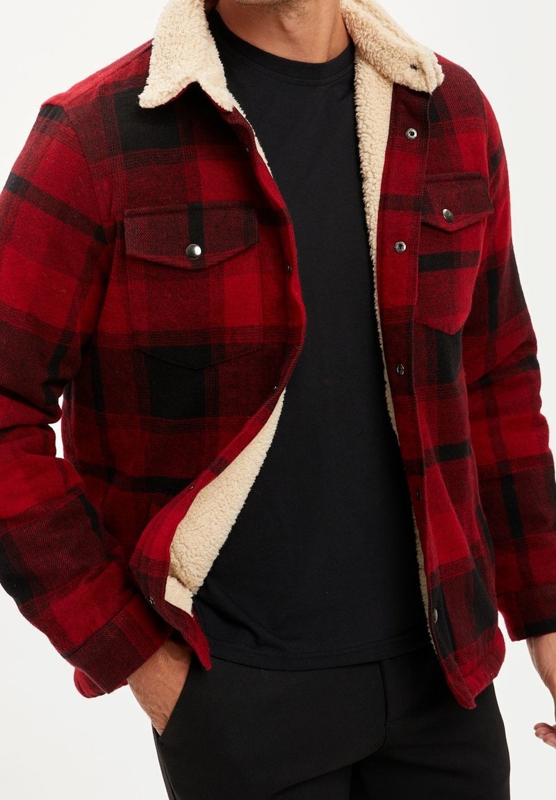 Man in een rood en zwart geruit jack met crème fleece voering, een zwart shirt en zwarte broeken, met zijn hand in zijn zak.