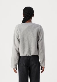 Lys grå, teksturert fleecegenser med rund hals, senkede skuldre og lange ermer. Kombineres med mørke jeans, minimal synlig detaljering.