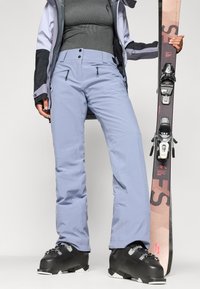 Pantaloni de schi de culoare albastru deschis, fabricati dintr-un material durabil, cu două buzunare cu fermoar, asociați cu cizme de schi negre și o schi cu legături.