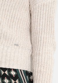 Suéter de punto beige con dobladillo acanalado y una pequeña etiqueta metálica "Hollister", combinado con pantalones de patrón negro, verde, rosa y beige.