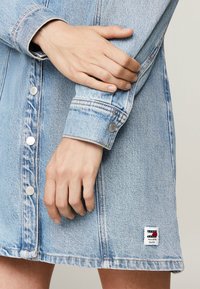 Light blue denim dress with button closure and silver snaps. Features a branded patch at the hem and visible stitching details.