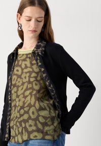 Zwarte jas met een kraag van luipaardprint, over een semi-doorzichtige, groen gepatterneerde top. Heeft een rits sluiting en een ontspannen pasvorm.