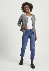 Giacca bomber trapuntata grigia con cerniera, indossata sopra una maglietta bianca, abbinata a jeans blu e stivaletti neri, su una persona con capelli ricci.