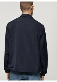Veste légère bleu marine à coupe décontractée, col montant et poignets élastiqués, présentant une texture lisse et un design minimaliste.