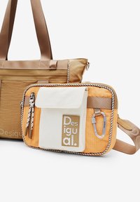 Dwie torby wyeksponowane: duża brązowa torba z grubymi uchwytami oraz mniejsza pomarańczowo-biała torba na ramię z logo i elementami hardware.