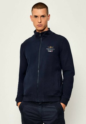 Giacca zip-up blu navy in morbido tessuto con colletto alto, con un design ricamato sul petto. Polsini e orlo a coste.