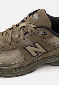 Baskets New Balance marron et olive avec panneaux en mesh et daim, lacets beige, bordures noires et logo "N" sur le côté.