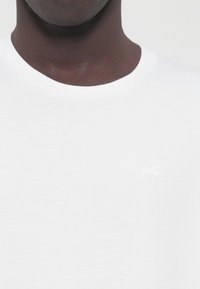 White cotton t-shirt with a round neckline and subtle logo detail in a lighter shade. Smooth texture, casual fit, suitable for everyday wear.