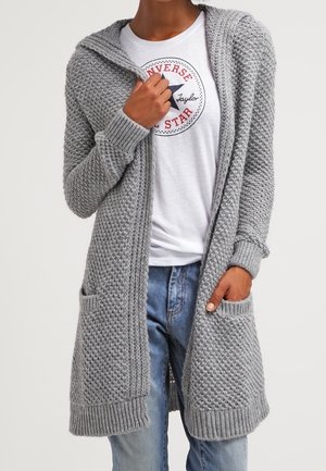 Personne portant un cardigan en maille grise texturée avec poches avant, sur un t-shirt blanc à motif Converse et un jean bleu.