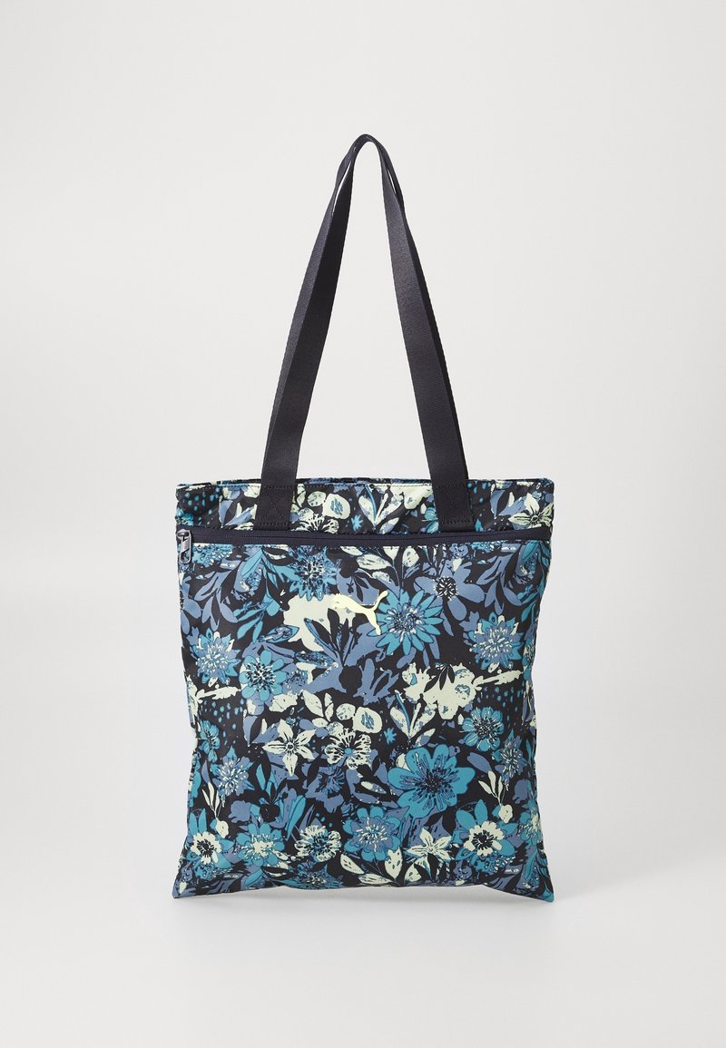 Sac fourre-tout à motif floral bleu et noir avec des anses foncées et une poche zippée à l'avant, suspendu devant un fond blanc uni.