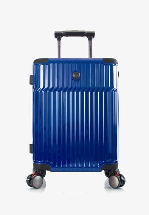 Maleta dura azul con ranuras verticales, asa negra extendida y cuatro ruedas giratorias dobles sobre fondo blanco.