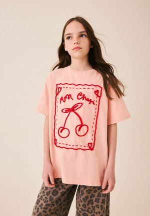 Dziewczyna z długimi ciemnymi włosami nosi różowy oversize t-shirt z czerwonym haftem wisienek oraz spodnie w panterkę, stojąc na beżowym tle.