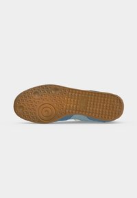 Semelle de chaussure en caoutchouc brun clair texturé comportant des motifs circulaires et triangulaires, avec un bord en tissu bleu visible au-dessus de la semelle.