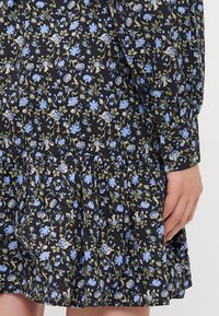 Abito di stoffa nero con stampa floreale di fiori blu e verdi. Gonna svasata con dettagli arricciati. Maniche lunghe con polsino.