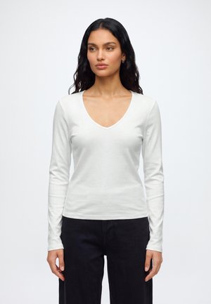 Femme aux longs cheveux foncés portant un chemisier blanc à col en V et manches longues et un pantalon noir, debout devant un fond clair uni.
