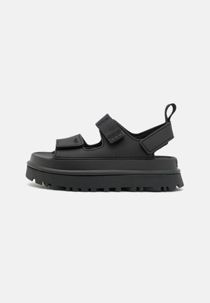 Sort sandal med en tyk platformssål, justerbare velcroremme og en struktureret overflade. Åben bagkonstruktion for åndbarhed og nem påføring.