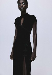 Vestido de terciopelo negro con cuello alto, mangas cortas y una abertura lateral, que presenta botones decorativos y detalles de costura curva.