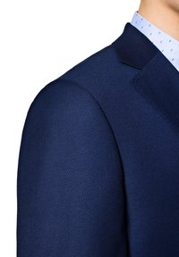 Granatowa marynarka z teksturowanej tkaniny, z wyraźnymi ramionami i klapami typu notch, w połączeniu z jasno-niebieską koszulą w małe wzory.