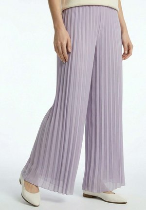 Femme portant un pantalon large plissé violet clair, assorti de chaussures blanches à petits talons, sur fond uni.