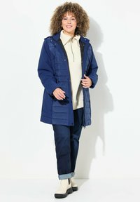 Cappotto puffer blu navy con chiusura a zip e automaticamente, dotato di una parte frontale trapuntata; abbinato a un leggero dolcevita e jeans con orlo arrotolato, e stivali color crema.