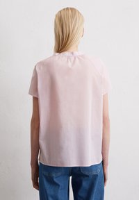 Kurzärmlige, hellrosa Bluse mit einem runden Ausschnitt, weichem Stoff und lockerer Passform. Verfügt über einen gerafften Detail auf der Rückseite. Kombiniert mit blauen Jeans.