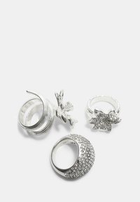 Fire sølvringe: en med strukturerede kurver, en med blomsterdesign og krystaller, en anden med en sløjfe, og en enkrusteret med små sten.